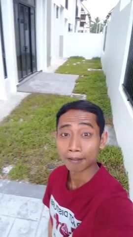 A medium shot captures a man with dark hair and brown skin wearing a maroon t-shirt. He is standing on a tiled walkway next to a patch of grass. Behind him, a modern building with white walls, black-framed windows, and a dark fence is visible. The man is looking directly at the camera with a neutral expression. The video appears to be a selfie as the camera is held by the man.