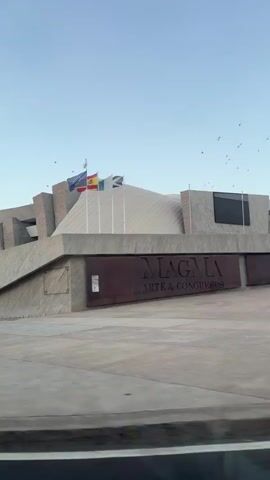 A video pans from left to right, capturing the exterior of a modern building with a distinctive architectural style. The building features geometric shapes, concrete facades, and a large, angled section covered in a lighter material. Flags are visible on poles in front of the building, and a sign reads "MAGMA ARTE & CONGRESOS." The scene is set against a clear blue sky with several birds flying overhead. The ground in front of the building is paved, and a road with yellow markings is visible at the bottom of the frame. The camera movement suggests it is being filmed from a moving vehicle.
