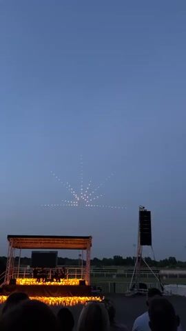 The video shows an outdoor evening concert at Kempton Park. A stage is set up with a band performing, and the stage is surrounded by numerous lit candles. In the sky above, a drone light show creates a radiant design resembling a sunburst. A crowd of people watches the performance from below. A large speaker tower stands to the side of the stage.