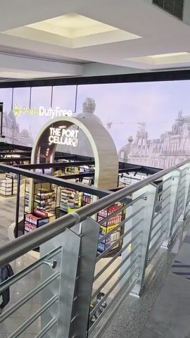 The scene is from a high vantage point looking down at a Porto Duty Free shop, called “The Port Cellar,” inside an airport. The shop is surrounded by glass display cases. A large arch-shaped wall and advertisement of Porto Duty Free is behind the shop. People walk through the shop and around it. A silver-colored railing is visible in the foreground. Overhead are several square recessed lights.