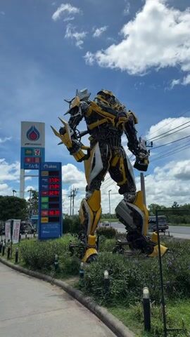 The video shows a large, yellow and black robot sculpture, resembling Bumblebee from Transformers, standing in front of a gas station. The robot is positioned on a grassy area with some purple flowers and small lights. Behind the robot, there is a tall blue and white sign with the PTT logo and a 7-Eleven logo. A price board displaying fuel prices is also visible on the sign, with numbers such as 37.99, 37.94, 38.02, 33.39, 36.92, 47.98. The gas station has several pumps and a covered area. In the background, cars are parked and driving, including a white van and several white cars. The sky is blue with scattered white clouds. The scene appears to be outdoors on a sunny day.