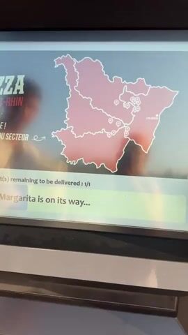 The video shows a close-up of a digital screen, likely a kiosk or display. The screen displays a map of a region in pink outline with several smaller icons, possibly representing delivery locations, scattered throughout. Text on the screen includes "KING OF PIZZA BAS-RHIN" and "MERCI POUR TA COMMANDE ! TOUTES NOS PLANQUES DU SECTEUR GEOLOCALISE SUR NOTRE APPLI". Below this, a status update reads "Product(s) remaining to be delivered : 1/1" and "Your Margarita is on its way...". The map also labels "STRASBOURG". In the background, two blurred figures, a man and a woman, are visible, their reflections also appearing on the screen. The lighting suggests it might be outdoors during the day, with a warm, possibly sunset, glow.