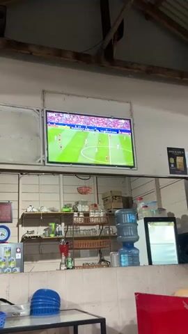 The video shows a television screen displaying a soccer match. The game appears to be a professional match, with players in uniform on a green field and spectators in the background. The scoreboard in the upper left corner shows "POLAND 0 - 0 ARGENTINA" and "76:35", indicating the match is likely from the 2022 FIFA World Cup. The television is mounted on a wall in what appears to be an indoor, casual setting, possibly a cafe or a home. Below the television, shelves are stocked with various items including glasses, bottles, and a water cooler. The lighting suggests it is daytime. There is also a sign with the text "ANNURARIKA" and an image of a coffee cup. The overall scene depicts people watching a sporting event.