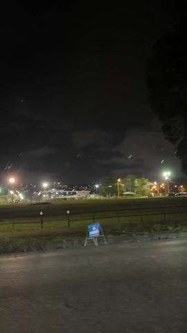 The video depicts a nighttime scene from a roadside, looking out onto a park or green space illuminated by streetlights. A "No Parking" sign is visible near the road. A car passes by the camera's field of view from right to left. In the background, there's a city skyline with numerous lights. The sky above is dark with some cloud cover. Trees are present on the left side of the frame, also illuminated by streetlights.