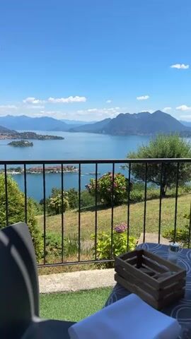 The video shows a scenic view of a lake with islands and mountains in the background. In the foreground, there is a balcony with a railing. A chair and a small table with a wooden crate and some white napkins are visible on the balcony. The lake is calm and blue, with several boats moving on its surface. Green trees and bushes, including a bush with pink and purple flowers, are scattered on the grassy slope leading down to the lake. The sky is clear and blue with a few white clouds. The overall impression is one of a peaceful and beautiful landscape on a sunny day.