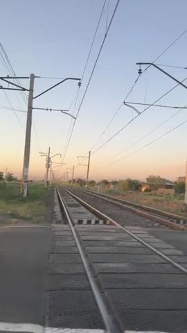 The video shows a railway crossing with overhead power lines and poles. The tracks stretch into the distance, disappearing into a hazy horizon. The sky is a clear, pale blue, transitioning to a warm orange hue near the horizon, suggesting either sunrise or sunset. Along the sides of the tracks, there is dry grass and scattered bushes. A paved road crosses the railway tracks, marked with a black and white striped crossing. On the right side of the road, there is a signal pole with a barrier arm, and a gray utility box. Further down the road, a white SUV is visible, driving across the frame from left to right. The camera appears to be positioned at a low angle, possibly from within a vehicle, looking towards the railway crossing.