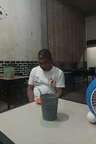 A man is sitting alone at a table in a sparsely furnished room. He is wearing a white t-shirt with writing and a logo. A small orange saucer with a glass and a spoon is in front of him. There is a gray, basket-weave textured holder on the table. The walls are a dull white with a black and white patterned stripe near the floor. In the background, there are dark tables and chairs, a striped curtain, and a doorway. There is a clock on the wall. The man is looking down at the objects on the table. The lighting is dim and the overall mood is subdued.