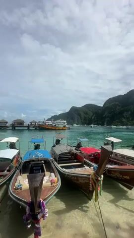 The media file depicts a picturesque scene at Ao Nang, Thailand, showcasing a fleet of traditional longtail boats docked in clear turquoise waters. The boats, varying in color from blue and red to natural wood, are lined up close to a sandy shore, their wooden prows adorned with colorful garlands and ribbons. Some boats have awnings providing shade.

In the background, a larger ferry or cruise ship is visible anchored further out in the bay. Beyond the water, lush green, mountainous islands rise dramatically, their peaks shrouded in a soft haze. The sky is partly cloudy, with white clouds scattered across a bright sky, suggesting it is daytime, likely late morning or early afternoon, with fair weather.

The scene evokes a sense of tropical tranquility and the promise of island adventures. The boats are clearly waiting to transport tourists, hinting at the bustling tourism industry of Ao Nang. The water is exceptionally clear, allowing the sandy bottom and the submerged parts of the boats to be visible.

Visible text includes boat registration numbers, such as "001" and "D9084", painted on the sides of some of the longtail boats. The overall impression is a classic Thai beach resort setting, abundant with natural beauty and activity.