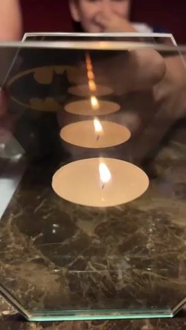 A close-up shot shows a row of five lit tea candles on a marble-patterned surface. A hexagonal mirror is placed above the candles, reflecting them and creating the illusion of a continuous line of light. The mirror has a partial image of the Batman logo visible on its surface. In the background, the blurred image of a young person is visible, looking downwards, with their hands partially obscuring their face, as if observing the reflection. The lighting is dim and appears to be from the candles themselves, with some ambient light in the background.