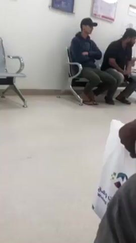The video shows the interior of what appears to be a waiting room. The room has a light-colored, speckled floor and white walls.  Two men are sitting in metal chairs in the background. The man on the left is wearing a dark hooded sweatshirt, green pants, a black baseball cap, and sandals. He has his arms crossed.  The man on the right is wearing a dark shirt and grey pants and appears to be looking at or interacting with something in his hands. A third metal chair is empty and to the left. There are framed posters or diagrams on the wall behind them. In the foreground, a person wearing a dark grey or black garment is holding a white shopping bag with branding on it.