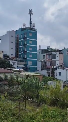A video displays a medium shot of a cluster of buildings under a cloudy sky. A tall, teal-colored building with a cellular tower on top is on the left. To its right are several multi-story residential buildings, varying in color and architectural style, some with balconies. In the foreground, lush green foliage and some thin bamboo-like plants are visible. The overall impression is of an urban or suburban landscape, possibly in Southeast Asia, given the building styles and vegetation.