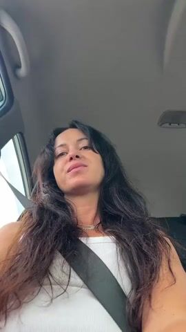 A woman with long, dark hair, wearing a white tank top and a seatbelt, is in the passenger seat of a car. She smiles at the camera, her head tilted slightly. The interior of the car is visible, as is a hint of the outside environment through the window, suggesting a daytime setting.