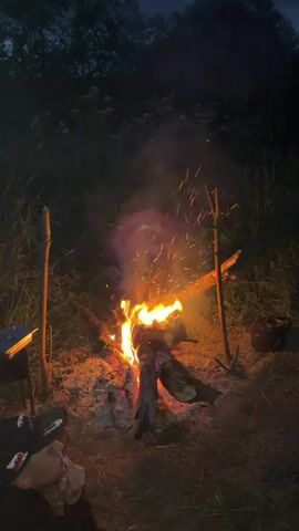 The video shows a nighttime scene outdoors with a campfire. In the foreground, a person wearing a black baseball cap is partially visible, looking to the left and holding something near their mouth. The campfire is in the center of the frame, with flames burning brightly and emitting sparks. There are logs burning in the fire, and ashes are visible on the ground. Two wooden poles are planted in the ground on either side of the fire, and a branch is resting across them, with another piece of wood supporting it. To the right of the fire, a dark pot or kettle is visible on the ground. In the background, trees and vegetation are dimly lit by the firelight, and the sky appears dark, indicating it is nighttime. The overall environment suggests a camping or outdoor gathering setting.