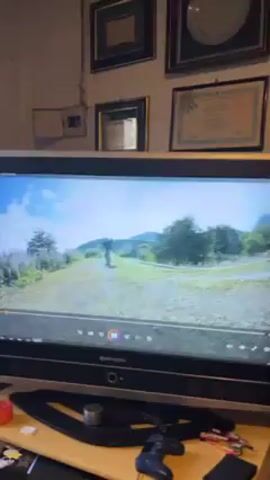 The video shows a person skateboarding in a video game on a Hitachi television. The game's graphics are of a natural environment with green hills, scattered trees, and a partly cloudy sky. The skateboarder, wearing casual clothing, navigates the terrain. Below the TV, there's a desk with a game controller, a small container, and some small red and white objects. On the wall behind the TV, a collection of framed pictures and certificates are displayed.
