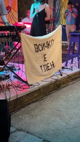 The video opens with a medium shot of a woman in a black dress singing into a microphone on a stage. To her left, a man with curly hair plays a keyboard. Behind them, a banner with black lettering in Cyrillic reads "ВСИЧКО Е ПЛЕН," which translates to "Everything is captured." The stage is decorated with colorful banners. To the right, another man is visible holding a phone. The lighting is dim, with pink and yellow stage lights illuminating the performers and equipment. The overall setting appears to be an outdoor performance venue.