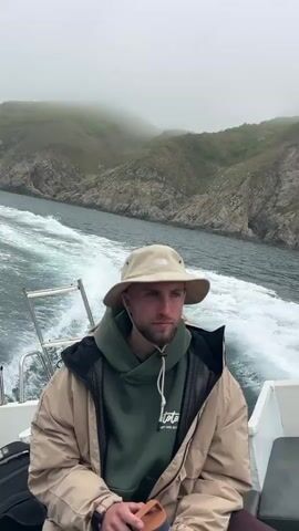 A man in a khaki bucket hat, green hoodie, and tan jacket sits on a white boat as it cuts through the water. He also wears maroon pants and holds a brown water bottle. The boat's wake is visible. Land covered in green vegetation and rocks is visible in the background, shrouded in fog. The camera slowly zooms out to reveal more of the boat and the seascape.