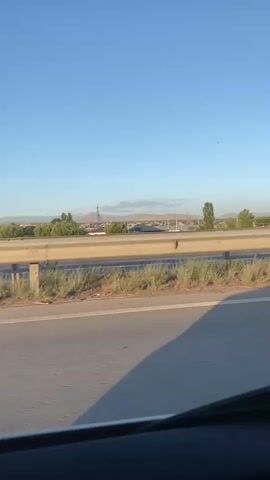 The video shows a view from a moving vehicle, likely a car, on a highway. The foreground is dominated by the road and a yellow metal guardrail. Beyond the guardrail, there are some dry grasses and shrubs. In the middle ground, there are trees and a low-lying landscape with some buildings, possibly a village or town, and a clear blue sky above. In the distance, there are mountains under a hazy sky. There are no people or explicit actions visible, other than the implied movement of the vehicle. The overall impression is a roadside scene during daylight.