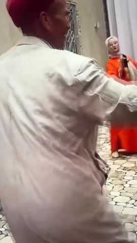 The video shows a group of people in an outdoor courtyard. The foreground is dominated by a person from behind, wearing a light-colored tunic and a red hat. This person is dancing or moving energetically. To the left, another person is playing a large drum. In the background, several women are visible, also dancing. The women are wearing colorful dresses, primarily orange and red. One woman on the far right is wearing a pink dress. The courtyard has a tiled floor and a wall with a metal-barred window on the left. A building with curtains and signage is visible in the background. The overall atmosphere appears to be a festive gathering or celebration.