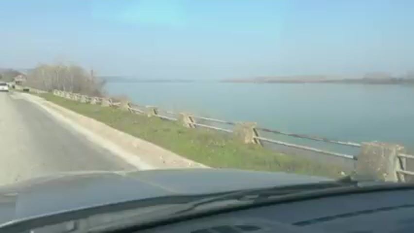 The video shows a view from the windshield of a vehicle driving along a road next to a river. The river is wide and calm, with trees lining the opposite bank. A low, weathered guardrail runs along the side of the road separating it from a grassy area that slopes down to the river. The sky is light blue and hazy. The road is asphalt and has a painted curb. The windshield wipers are visible at the bottom of the frame.