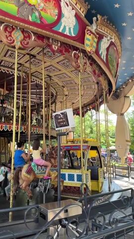 The video shows a carousel in operation. The carousel is elaborately decorated with painted murals and gilded accents. The murals depict cartoon characters and medieval-themed scenes. The carousel features various animals for riders, including horses and what appears to be a truck or carriage. Several people are visible riding the carousel. One person in a blue shirt and denim shorts is seated on a horse, while another person in a pink shirt is on a different horse. In the background, more people and carousel animals are visible. The setting appears to be an amusement park or fairground, with trees and fencing visible beyond the carousel. The sky is blue with white clouds, suggesting daytime. The overall mood seems joyful and lighthearted, as is typical of carousel rides. There is some text visible on a white box mounted on a pole, but it is too blurry to read. The location context provided is Castelnuovo del Garda, Italy, which is home to Gardaland, an amusement park known for its elaborate rides.