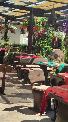 The video shows an outdoor seating area of a restaurant or cafe, possibly in a village or rural setting. The tables and benches are made of rough, dark wood, with some tables draped in red and white striped cloths. Hanging baskets filled with pink and purple flowers are suspended from a wooden overhead structure, which is covered with a light-colored fabric canopy, allowing dappled sunlight to filter through. Greenery and plants are abundant throughout the scene. The atmosphere appears to be peaceful and inviting.