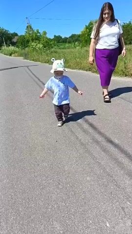 A young child wearing a blue and white striped button-down shirt, dark pants, and a beige hat with frog ears is walking down a paved road. A woman in a white t-shirt and purple skirt is walking behind the child. The child has a pacifier in their mouth and is holding a red lollipop in their right hand. The scene is outdoors on a sunny day, with green grass and trees on either side of the road. The child's shadow is cast on the road.