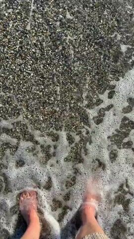 A first-person perspective video shows feet stepping into gentle ocean waves on a pebble beach. The water is clear and the sun is shining, creating a shimmering effect on the surface. The waves wash over the feet and ankles, creating foam and ripples. The pebbles on the beach are small and dark, mixed with some lighter colored ones. The video captures the sensation of being at the beach and enjoying the cool water on a warm day.