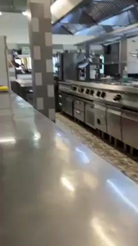 The camera slowly pans across a large, well-lit, industrial kitchen. The space features extensive stainless steel surfaces, including countertops, appliances, and ventilation systems. The appliances consist of multiple ovens, ranges, and refrigerators. The floor is a patterned tile. A pillar with a grey and white tiled design stands in the middle of the kitchen. People are faintly visible working in the background.