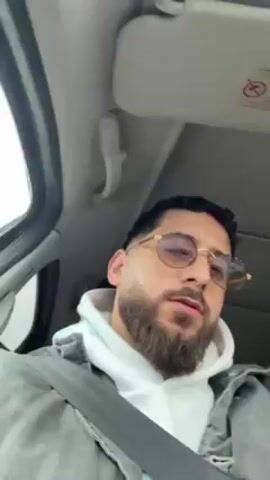 The video shows a bearded man with sunglasses sitting in a car, wearing a white hoodie and a gray denim jacket. He gestures with his hand while listening to music, appearing to mouth the words or sing along. The car's interior is visible, including the sun visor, seatbelt, and side mirror. The scene is captured from a low angle, focusing on the man's upper body.