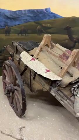 A close-up shot of an antique wooden cart filled with assorted items. In the background, a mural depicts a desert landscape with camels, yurts, and people. The cart itself is laden with burlap sacks, a rope coiled on top, and what appear to be textiles or rugs. A large, rough-hewn wooden wheel is visible on the left side of the cart. To the right of the cart, a stone grinding mill with a wooden handle rests on a base of rocks, suggesting a historical or cultural exhibit. The lighting appears to be artificial, illuminating the scene within an indoor setting.