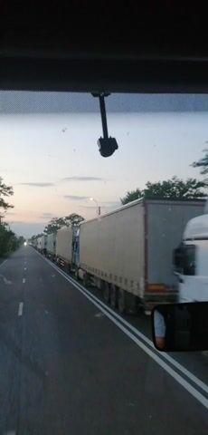 The video, filmed from inside a vehicle, shows a line of semi-trucks parked on the side of a road. The sky is a pale blue with hints of pink and orange, suggesting it is either dawn or dusk. The trucks are all white with some displaying logos or text. The road has white lane markings. A rear-view mirror is visible, reflecting a person's face and a portion of the interior of the vehicle from which the video is taken. There is a black object mounted on a pole on the left side of the frame. The overall impression is a quiet, still moment on a highway.