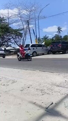 The video shows a street scene under a clear blue sky with scattered clouds. In the foreground, there is a dirt or gravel surface with some scattered debris. A paved road runs through the scene. Several motorcycles are parked on the side of the road, and some are in motion. One woman wearing a pink headscarf and pink clothing is riding a blue and white motorcycle. She is the central focus of the initial frames. In the background, there is a building with a red and white canopy, possibly a gas station, with flags. There are also trees and utility poles visible. The overall atmosphere is that of a bright, sunny day in a somewhat rural or suburban area with significant motorcycle traffic.