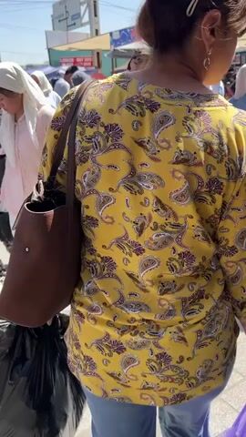 The video captures a bustling outdoor market scene on a sunny day. A woman in a yellow paisley shirt is in the foreground, facing away from the camera. In the background, several people are walking and interacting. A woman in a blue shirt and hijab is talking to a man wearing a bucket hat and a backpack. Another man in a red shirt and denim shorts stands to the right. The environment appears to be a street market with stalls and various people browsing. The ground is paved with cobblestones, and shadows are cast by the sunlight.