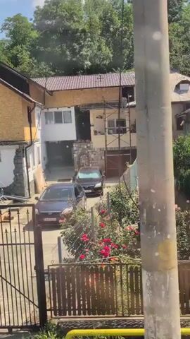 The video pans across a residential area with several houses, some appearing to be under construction or renovation. In the foreground, a black metal gate and fence enclose a driveway where two dark-colored sedans are parked. Beyond the gate, two houses are visible; one has a yellow stucco facade with a dark-trimmed window and a small balcony, while the other is partially obscured by trees and scaffolding. Further in the distance, more houses and lush green trees can be seen under a bright, partly cloudy sky. The camera moves slightly, revealing a closer view of the yellow house, showing a staircase leading to a dark doorway. Another house, a lighter color, is situated next to it. The video continues to pan, showcasing a white building with a dark roof, and then a wider view of the neighborhood with more homes nestled among trees on a gentle slope. A fence, partially covered in greenery, separates the properties. The final shots offer a broader perspective of the houses and their surroundings, with a clear blue sky overhead.