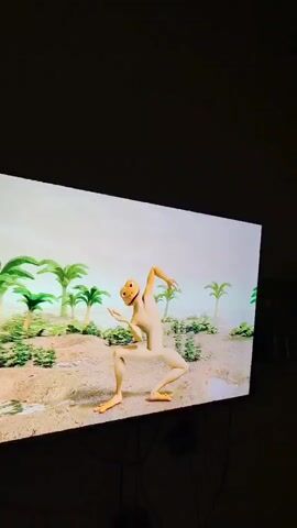 A television screen displays an animated frog character dancing in a desert-like environment with palm trees. In the foreground, three children are watching the television. A girl in a pink shirt is in the middle, clapping her hands and smiling. To her right, a boy in a blue and red shirt is looking at the screen. Another child with dark hair and a white shirt is on the left, with their back to the camera. The children appear to be in a room with a tiled floor and neutral-colored walls.
