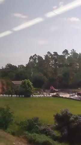 The video shows a view from a moving vehicle, likely a train, looking out a window. The scene outside is a landscape with trees, some buildings, and green fields. There is a reflective element on the left side of the frame, possibly a pillar or a part of the vehicle's interior, which obscures portions of the view and reflects the sky and surroundings. The sky is partly cloudy. The overall impression is of a journey through a rural or suburban area on a bright day.