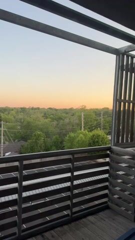 The video shows a view from a balcony overlooking a green, leafy landscape, likely trees, under a sky with a soft gradient of colors suggesting dawn or dusk. The balcony has a metal railing with horizontal slats and a partial metal and wooden structure above. Telephone poles and wires are visible among the trees.