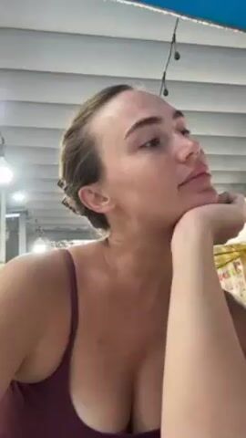 A woman with her hair pulled back is looking at the camera, resting her head on her hand. She is wearing a maroon tank top. A child is standing next to her. The area appears to be an outdoor dining space with white ceiling slats and string lights overhead.