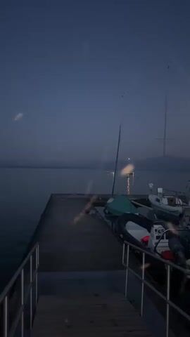The video shows a dock on a body of water at dusk. Several boats are docked, including a sailboat with a tall mast and a smaller inflatable boat. The sky is a deep blue, with the moon visible as a bright disc. The water is calm, reflecting the sky and the lights of a distant boat. There are two people standing on the dock in the background, seemingly observing the scene.