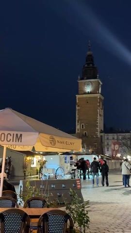 The video shows a night scene in a European city square. Several outdoor cafes are set up with tables and chairs under umbrellas. People are seated at some of the tables. There is a tower clock illuminated in the background. Horse-drawn carriages are parked nearby. People are walking around the square. One person walks away from the camera and into the distance. A gas heater is visible in the cafe area. The sky is a deep blue.