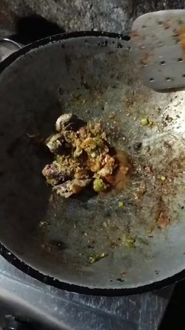 A close-up video shows food being cooked in a wok. A metal spatula is used to stir the food, which appears to be a mixture of fish, vegetables, and spices. The wok is placed on a gas stove, and the background is dimly lit, suggesting the video was taken at night or in a dark setting. The food appears to be in the process of being fried or stir-fried.