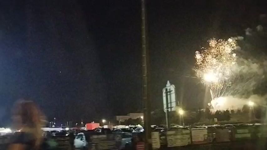 This video captures a nighttime scene with fireworks and a carnival ride. In the foreground, two individuals walk from left to right across the frame. Behind them, a parking lot filled with cars is visible. A tall, brightly lit carnival ride, resembling a swing ride, stands on the left side of the image. On the right side, fireworks are exploding in the dark sky, illuminating the scene with bursts of light and smoke. Traffic lights and streetlights can also be seen in the background. The overall atmosphere suggests a festive event like a fair or a celebration.