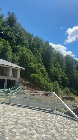 The video presents a landscape view on a sunny day. A gray and white brick-paved area is in the foreground, bordered by a white fence. Beyond the fence is an area of exposed dirt with some wooden planks lying on it. The middle ground features a steep hillside densely covered with green trees. In the background, there is a bright blue sky with scattered white clouds. A building under construction is visible to the left side, partially obscured. The video appears to be taken from a stationary position with no visible people or movement.