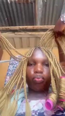 A woman is the primary focus of this close-up video. Her face is centered in the frame as she puckers her lips and poses for the camera. She is wearing light makeup and has blond braided hair, with some sections rolled into pink curlers. Two pairs of dark-skinned hands are visible around her head, holding and arranging the braids. One hand is using a pink brush to style the hair. The video appears to be taken outdoors, judging by the corrugated metal roofing overhead. A poster or photograph is visible in the background.