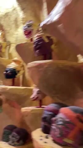 The video shows a close-up panning shot of an indoor display featuring several decorative masks or puppets displayed on tiered, rock-like shelves. The masks are of various colors and designs, some with elaborate headdresses. The lighting is dramatic, with pink and purple hues illuminating the scene, creating a theatrical or exhibition-like atmosphere. The background appears to be a textured, rocky wall, possibly part of a themed exhibit. No people are clearly visible in the frame.