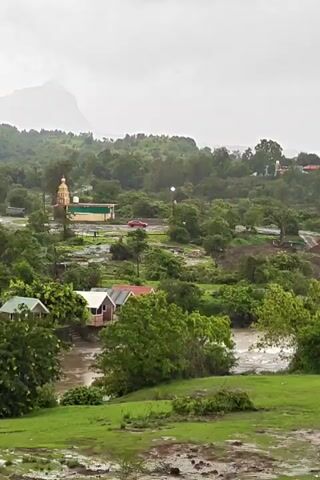 A wide, panning shot captures a lush, green landscape under an overcast sky. In the mid-ground, several small, colorful houses with red roofs are nestled near a flowing river. Further back, a golden-domed temple stands prominently, surrounded by trees. A red car is visible on a road near the temple. In the distance, a large, mist-shrouded mountain looms. The ground in the foreground is covered in wet grass and patches of mud, indicating recent rainfall. The overall impression is one of a serene, somewhat damp, rural environment.