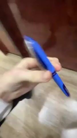 A pale-skinned person wearing a dark suit and a white cuff on their left wrist is holding a blue pen and twirling it between their fingers. The person is standing on a light-colored wooden floor near a dark brown wooden door. The video is slightly shaky.