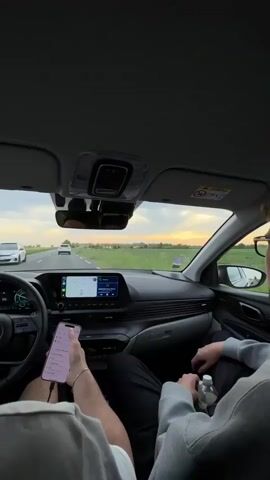 The interior of a car is shown during a daytime drive.  Two people are visible in the front seats. The driver is wearing a light-colored sleeveless shirt and has a phone in his hand connected with a cord. He appears to be looking at the phone. The passenger is wearing a gray hoodie and black pants, holding a water bottle. The passenger adjusts the car's touchscreen display. The car is driving on a highway with fields visible on either side. Other cars are on the road, and the sky is a mix of clouds and patches of sunlight. The car's dashboard and steering wheel are visible, including the speedometer showing a speed around 80-85.