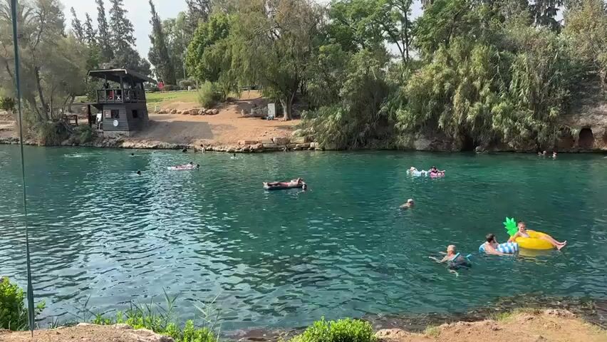 The video shows a body of turquoise water with several people swimming and floating on inflatables. The water is surrounded by a rocky embankment with lush green trees and vegetation. On the left side of the frame, there is a wooden structure with a roof and a small, enclosed area. In the background, more trees and a cloudy sky are visible. People are scattered throughout the water, some are swimming, while others are lounging on various inflatable objects, including a pink flamingo, a yellow pineapple, and a black raft. One person is in a kayak. The overall scene depicts a recreational area, likely a natural spring or a designated swimming spot, on a clear day.