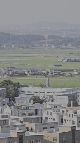 A panoramic, distant view of a semi-urban landscape. In the foreground, several multi-story buildings are visible, characterized by their modern, angular architecture and neutral color palettes of white, grey, and beige. Some buildings feature balconies and air conditioning units. Behind these buildings, a vast expanse of green fields, likely agricultural land, stretches into the distance. Scattered across the fields are a few scattered buildings, some appearing to be houses and others larger industrial or institutional structures. Further in the background, rolling hills form a natural backdrop, with some evidence of human development, such as roads and power lines, visible on their slopes. The sky is hazy and overcast, contributing to a muted color palette and a soft, diffused light across the entire scene.